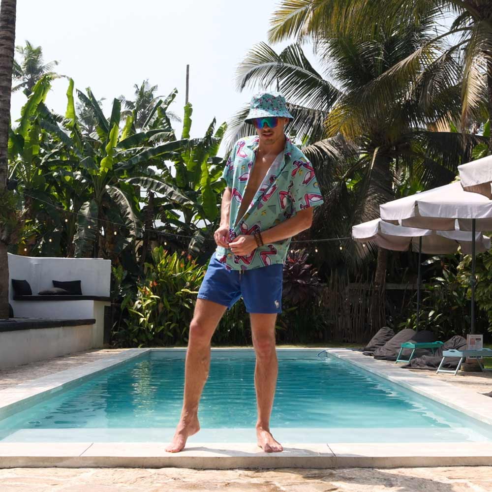 Wearing the Flamingo Fandango Shirt, blue shorts, and a sun hat, a man stands by a pool bordered with tropical plants and palms; lounge chairs and umbrellas sit in the background.