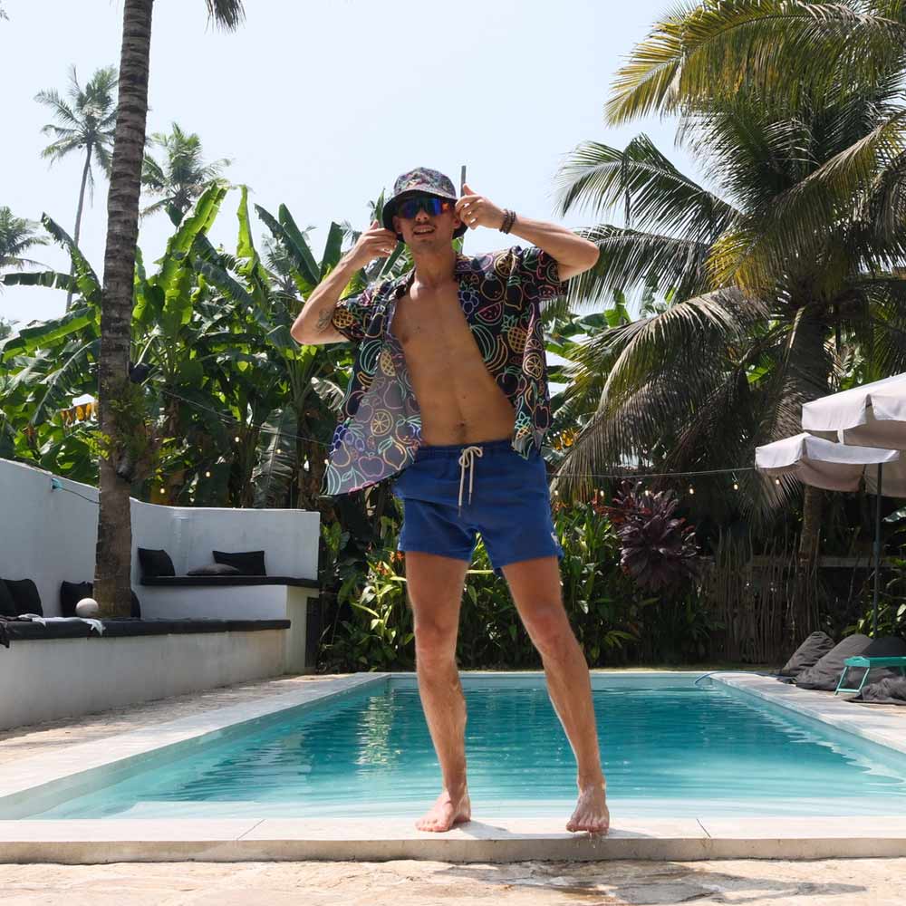 A man in blue swim shorts, a bucket hat, and the Fluorescent Fruit Fiesta Shirt poses playfully by a sunlit pool, surrounded by palm trees and lush greenery.