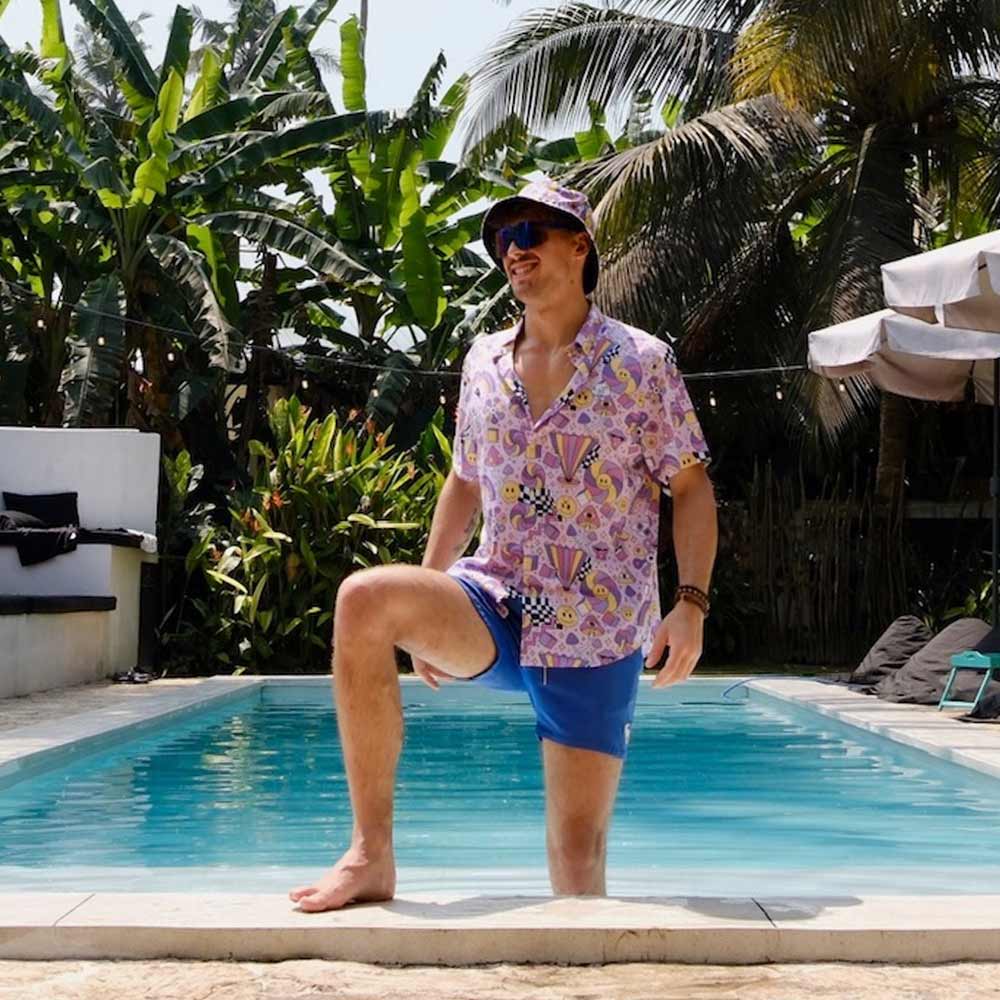 A man wearing a Hippie Shirt, blue shorts, and a bucket hat stands by a pool with one knee up, surrounded by lush tropical plants and palm trees.