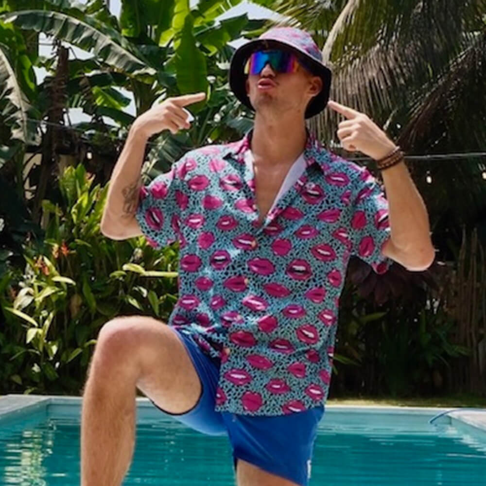 A man in a colorful shirt, blue shorts, and reflective sunglasses points at his Leopard Kiss Bad Bucket Hat while posing poolside. Lush tropical plants and palm trees create a vibrant backdrop.