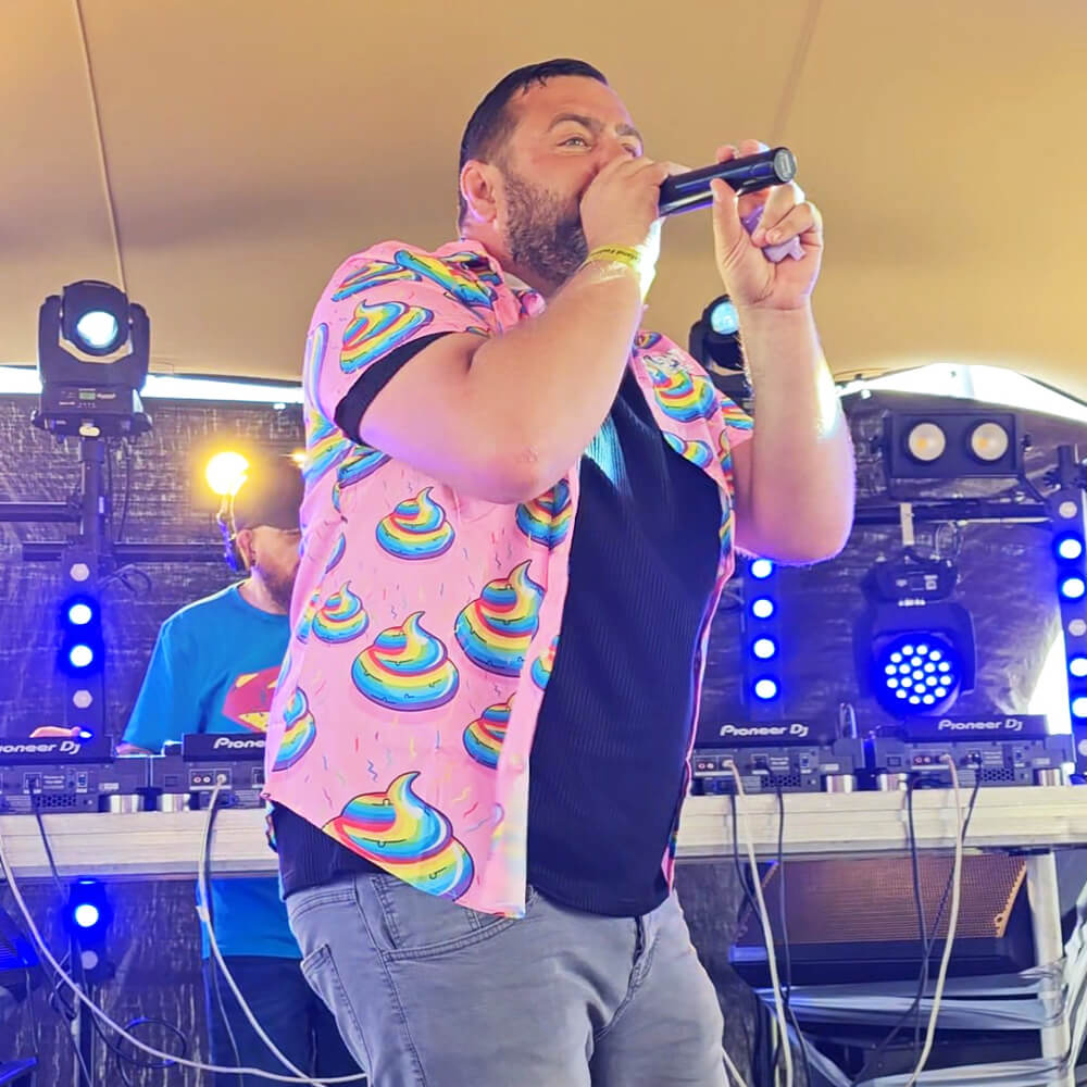 A man wearing a colourful shit shirt with rainbow swirl designs sings into a microphone on an outdoor stage, with DJ equipment and a person in a blue shirt in the background. Bright stage lights are visible.