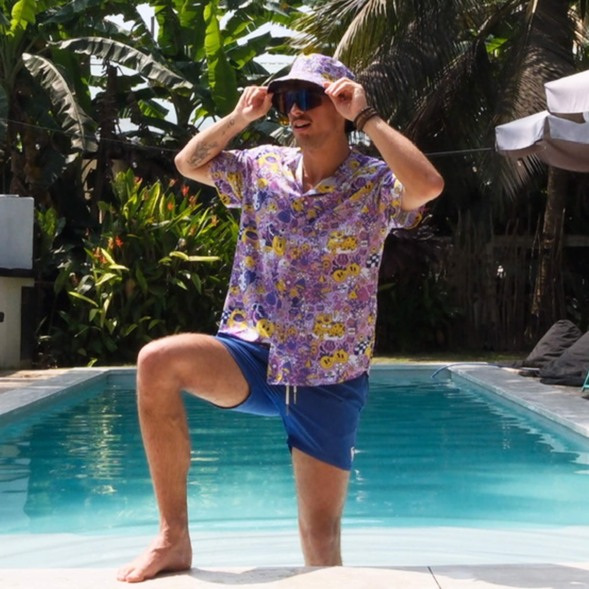 Wearing a Magical Psychedelic Shirt and matching bucket hat, a person in blue shorts poses confidently by a pool, surrounded by lush plants and palm trees on a sunny day.