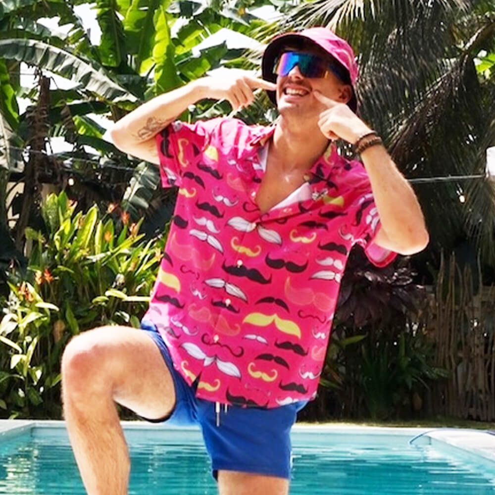 A smiling person in a pink Movember Shirt with mustache patterns, pink bucket hat, and sunglasses kneels poolside, playfully pointing to their mustache amid lush tropical plants.