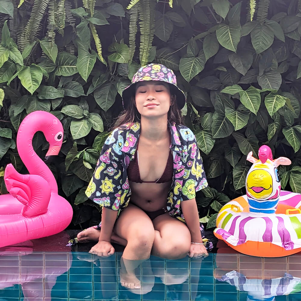 Woman wearing a mushroom shit shirt sitting by a pool with an inflatable flamingo, surrounded by green foliage.