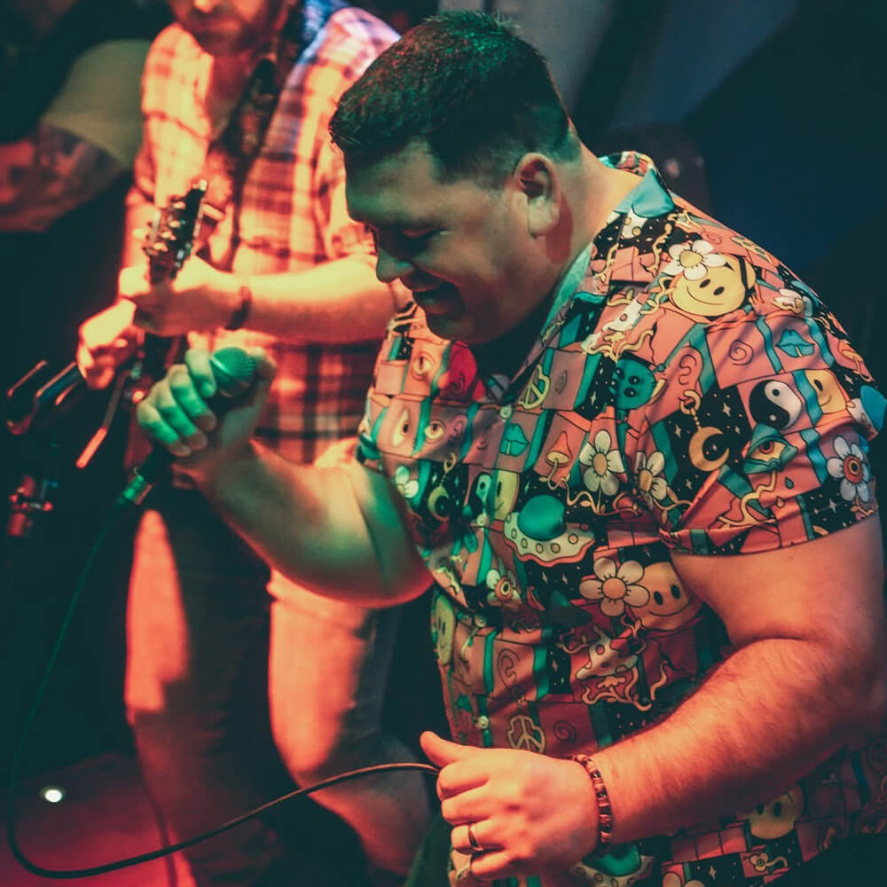 A man in a colourful, cartoon-patterned shit shirt passionately sings into a microphone on stage, while a guitarist plays in the background under vibrant lighting.