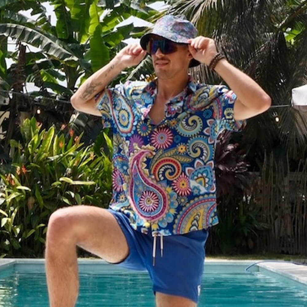 A man smiles and adjusts his Psychedelic Explorer Bucket Hat while posing by a pool surrounded by lush tropical plants. He wears a colorful patterned shirt and blue shorts, enjoying an outdoor setting.