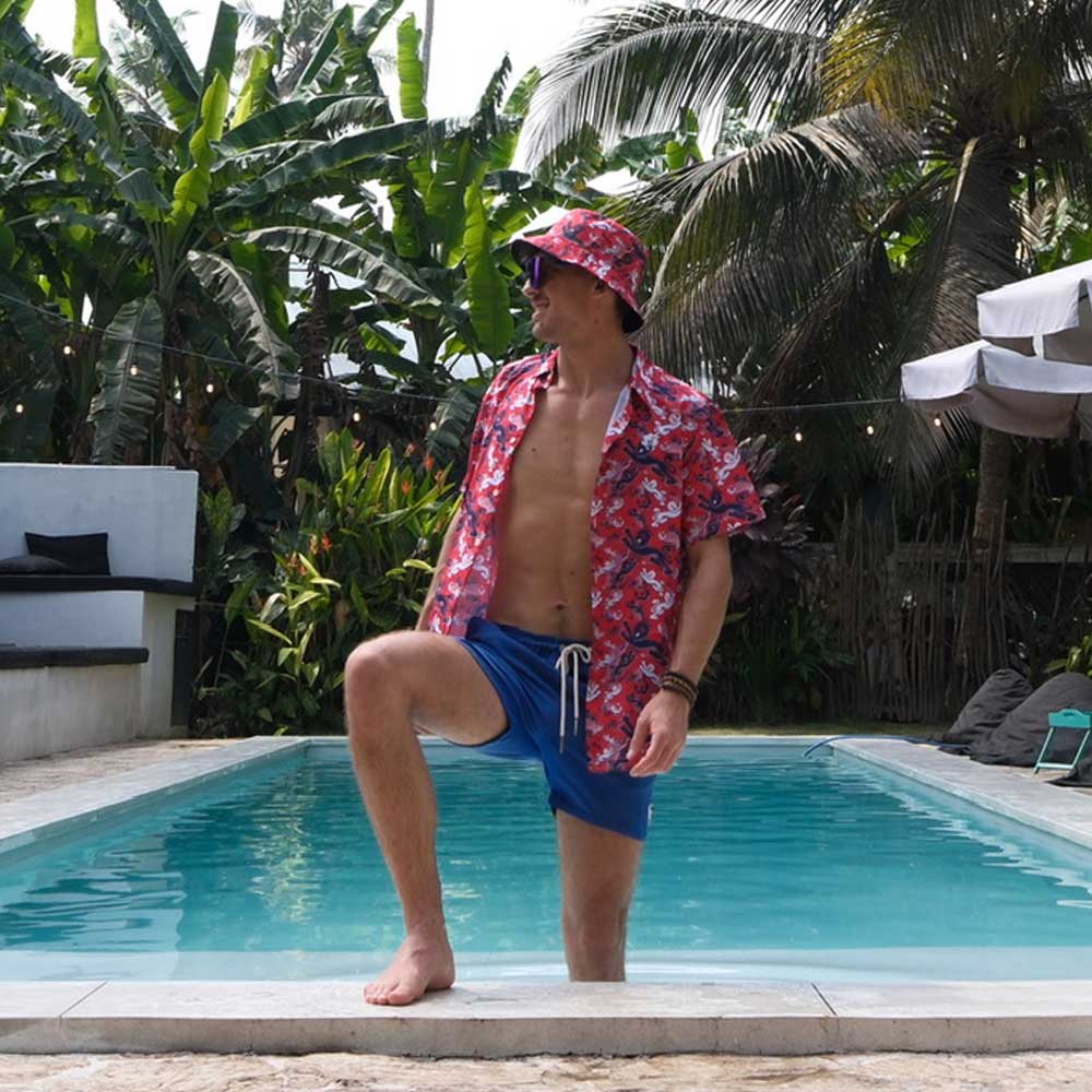 Wearing a Red England FC Hawaiian Shirt, blue shorts, and a matching hat, a person poses with one foot on the pools edge amid lush tropical plants and palm trees.