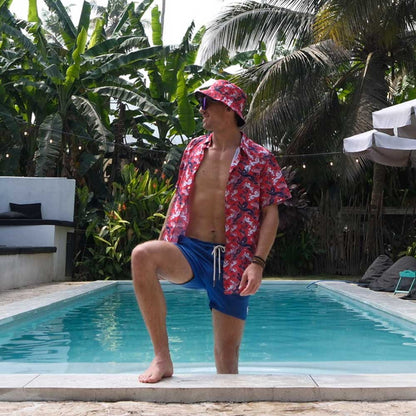 Wearing a Red England FC Hawaiian Shirt, blue shorts, and a matching hat, a person poses with one foot on the pools edge amid lush tropical plants and palm trees.