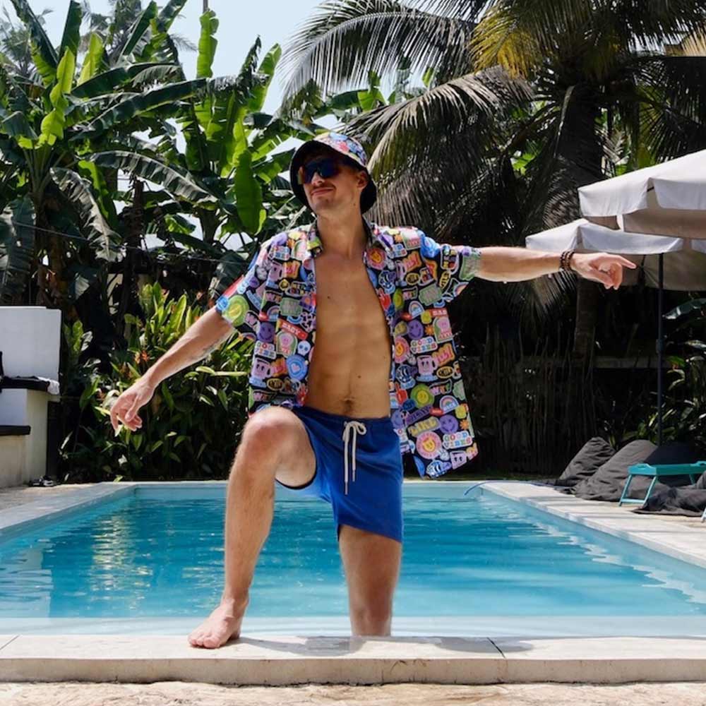 A man wearing the Retro Sticker Shirt, blue swim trunks, and a bucket hat poses poolside with one knee up and arms wide. Palm trees and umbrellas are visible in the sunny background.