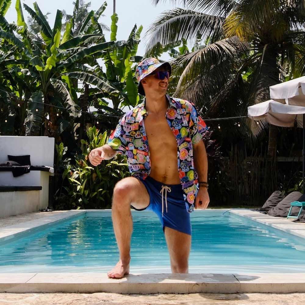 A man in a Retro Sticker Shirt, blue swim shorts, bucket hat, and sunglasses poses with one knee up by a pool, surrounded by tropical plants and palm trees.