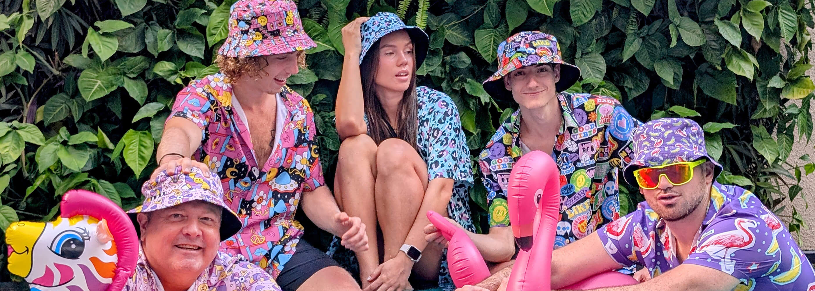 Five people in colourful, cartoon-patterned Shit Shirt Club outfits and matching bucket hats sit together with inflatable toys, including a flamingo and unicorn, in front of lush green plants. They appear relaxed and playful.