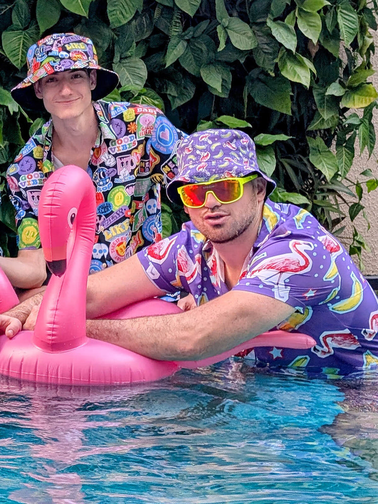 Two men in colourful shit shirts and matching bucket hats pose in a pool. One leans on a pink inflatable flamingo while wearing neon sunglasses. Green leaves are visible in the background.