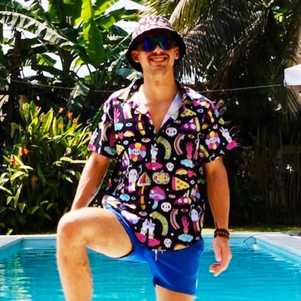 A smiling man in a colorful patterned shirt, blue shorts, and the Shroom Bucket Hat stands poolside amid lush tropical plants and trees on a sunny day.