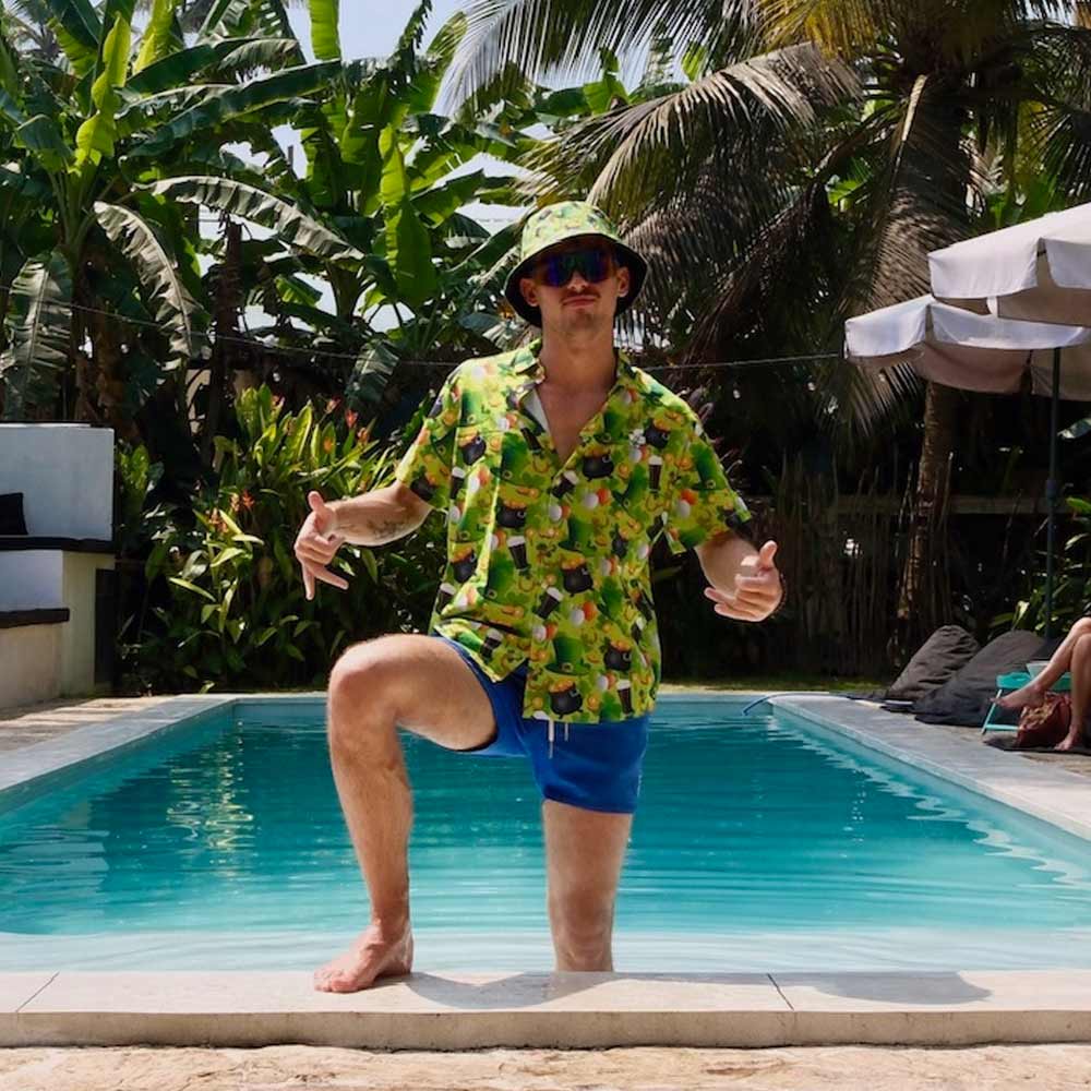 A man in a St Patricks Day Shirt, blue shorts, sunglasses, and a bucket hat poses poolside with one knee up and both thumbs up. Lush tropical plants and palm trees fill the background.