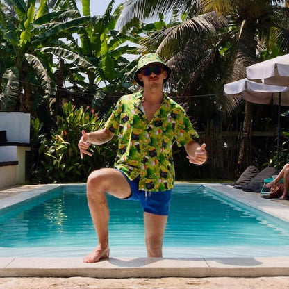A man in a St Patricks Day Shirt, blue shorts, sunglasses, and a bucket hat poses poolside with one knee up and both thumbs up. Lush tropical plants and palm trees fill the background.