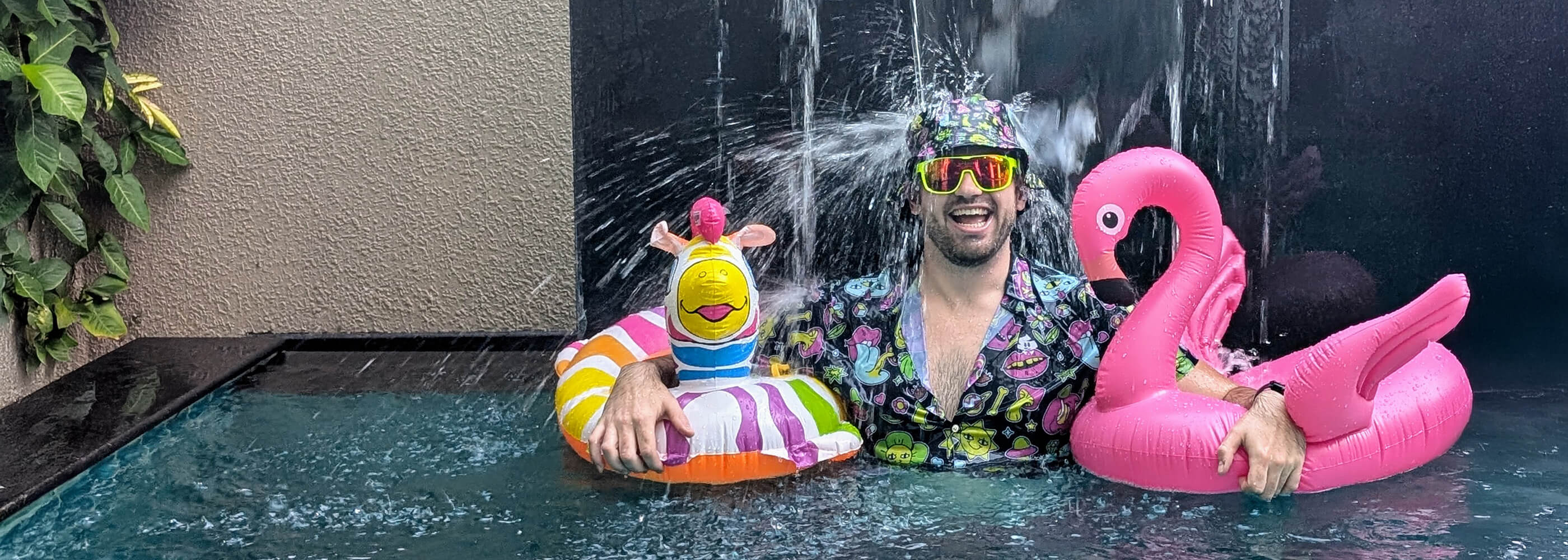 A man in a colourful shit shirt and neon sunglasses stands in a pool under a waterfall, smiling and holding inflatable floaties shaped like a pink flamingo and a rainbow unicorn. Green leaves hang on a nearby wall.