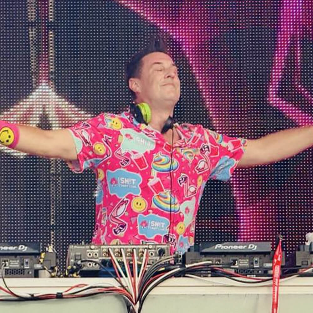 A DJ wearing the Badass Shirt, featuring a vibrant flamingo print, spreads his arms wide on stage with green headphones, DJ equipment, and bright LED lights in the background.