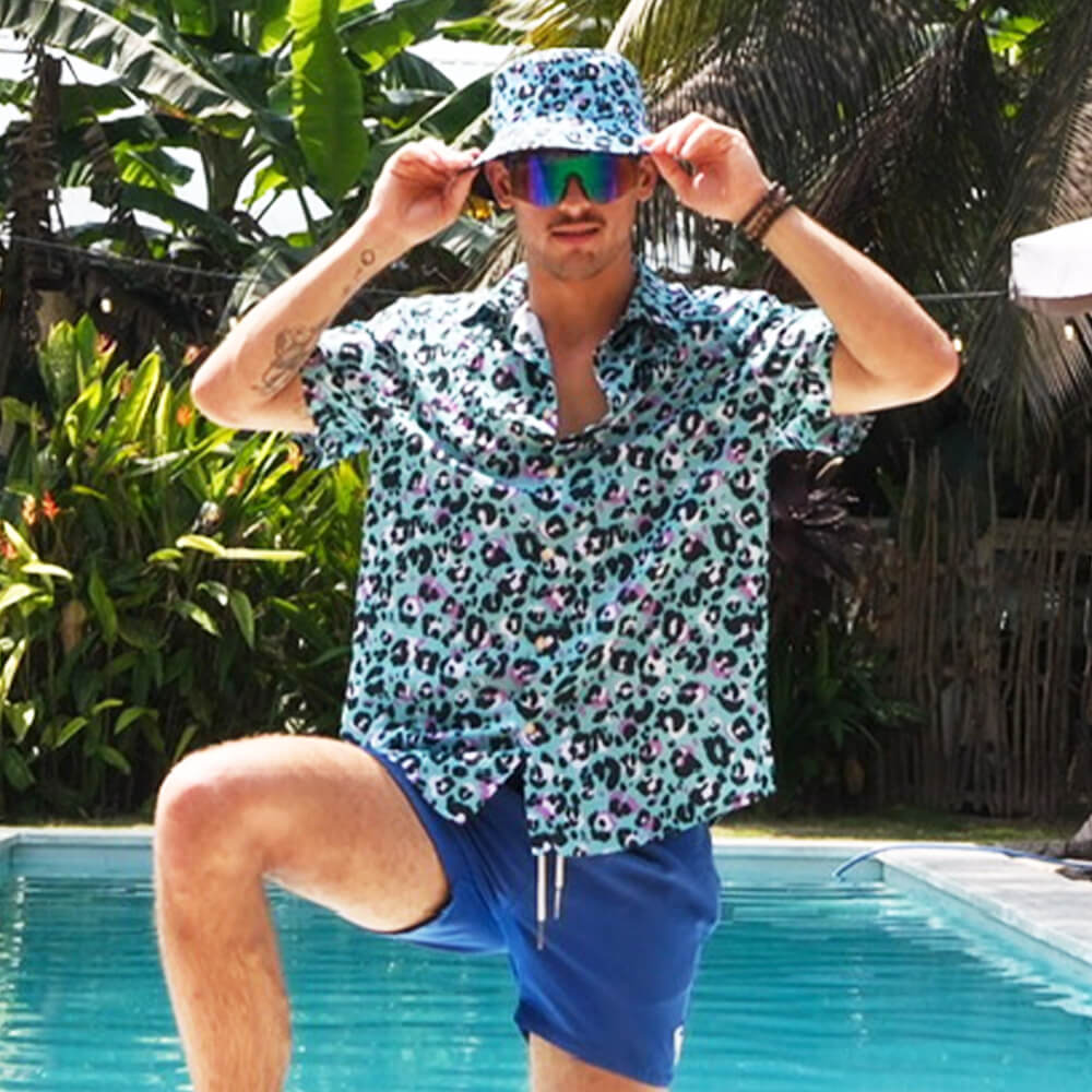 By the pool amid lush tropical plants, a man showcases bold style in the Turquoise Tempest Leopard Print Shirt, paired with a matching bucket hat, blue shorts, and reflective sunglasses.