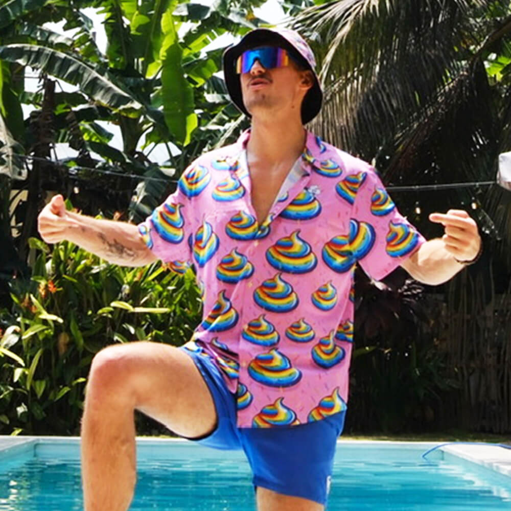By a pool, someone in blue shorts, sunglasses, and a pink shirt with rainbow poop emojis playfully poses wearing the Unicorn Poo Bucket Hat. Lush tropical plants fill the background.
