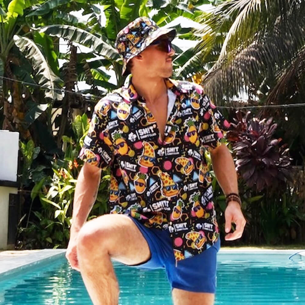 A man in the Wanna Pizza This Shirt and a bucket hat with cartoon graphics stands by a pool in blue shorts and sunglasses, surrounded by lush tropical plants.