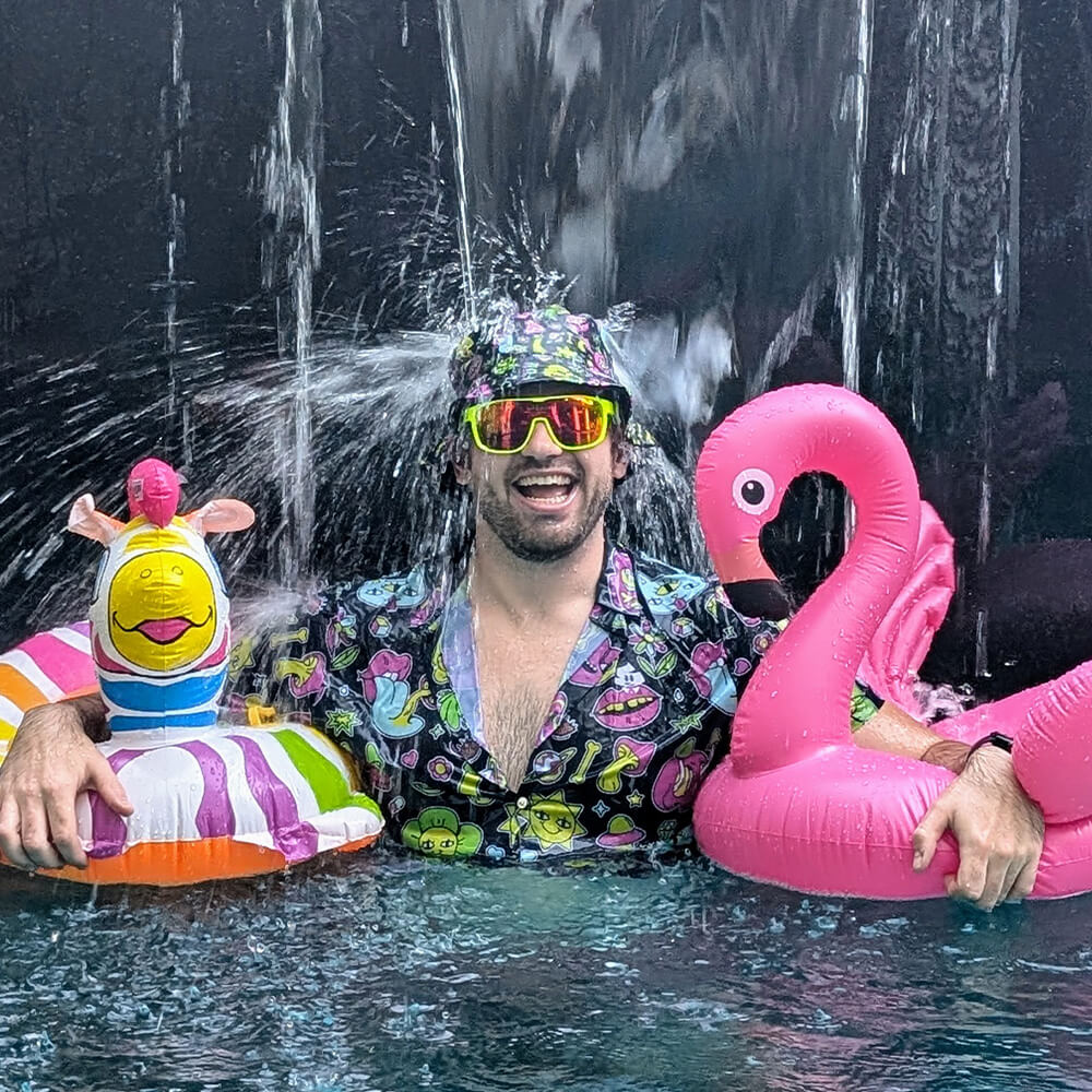 A smiling man wears Send It Shades, a patterned shirt, and matching hat while standing in a pool, holding an inflatable flamingo and a giraffe float, with splashing water behind him.