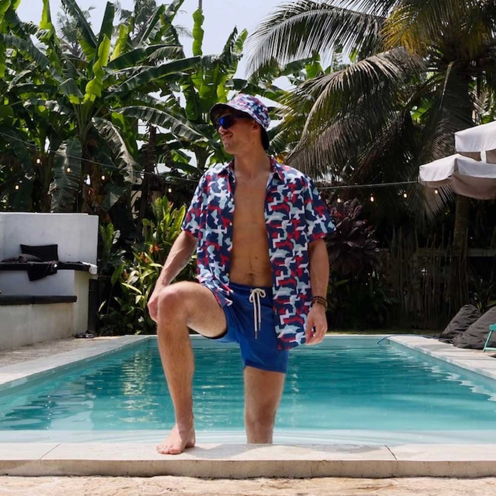 A man in a Combat2Coffee Army Camo Shirt, with one knee on a pool edge, is surrounded by lush tropical plants and palm trees—promoting mental health awareness in a vibrant setting.