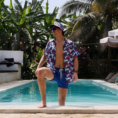 A man in a Combat2Coffee Army Camo Shirt, with one knee on a pool edge, is surrounded by lush tropical plants and palm trees—promoting mental health awareness in a vibrant setting.