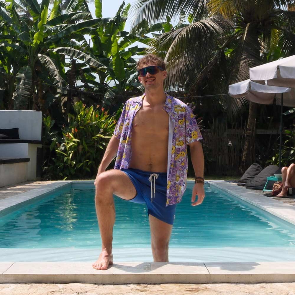 A man in blue swim shorts, sunglasses, and a Magical Psychedelic Shirt stands with one leg on the poolside and the other in the water, surrounded by palm trees and tropical plants on a sunny day.