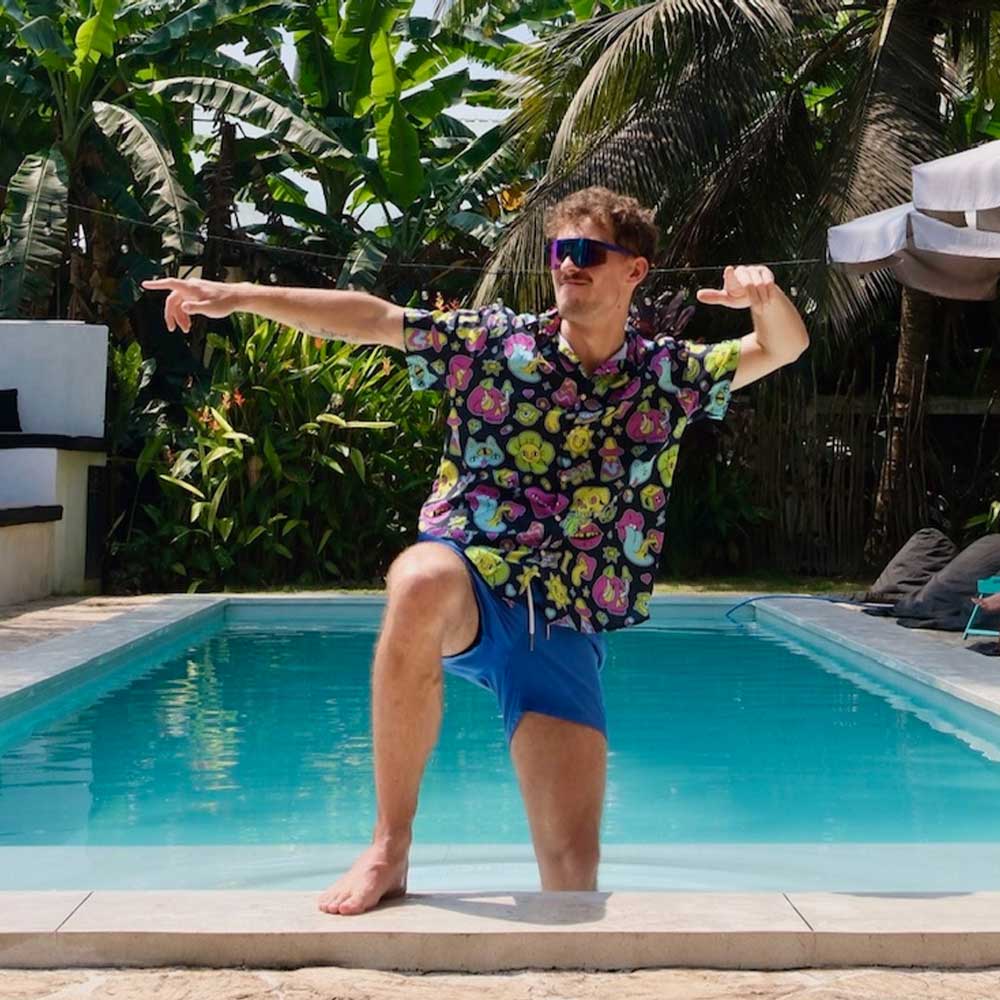 A man in blue shorts and a Black Out Mushroom Shirt poses playfully by the pool, one knee up and both arms outstretched. Sunglasses and lush tropical plants complete his bold festival fashion vibe.