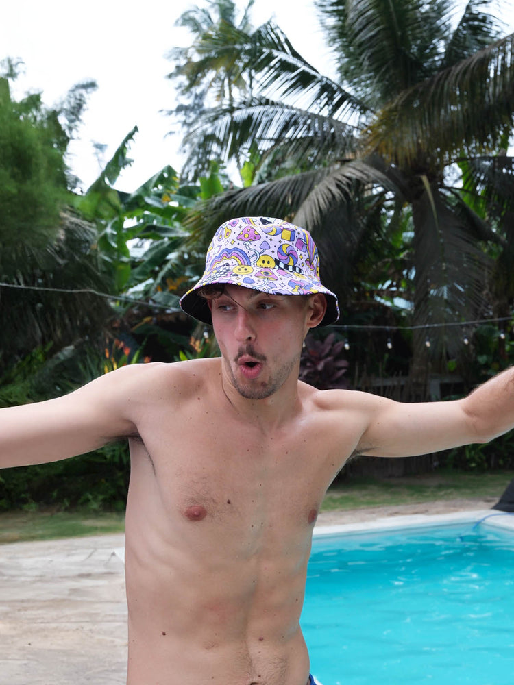 A shirtless man wearing a colorful bucket hat stands by a swimming pool, arms outstretched and making a playful surprised expression. Lush green trees and plants surround the outdoor pool area.