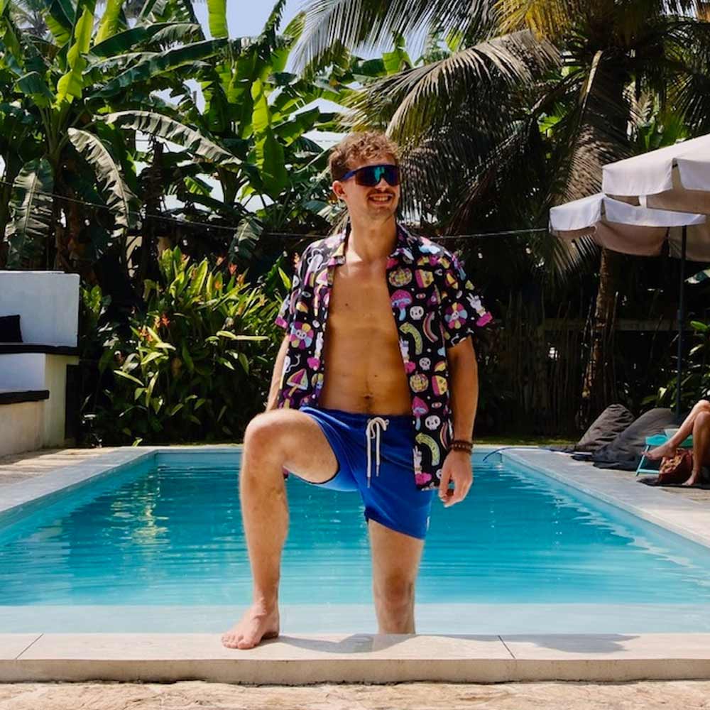 A man in blue swim trunks and a Shroom Shirt stands confidently at the pool’s edge, one foot on the ledge. He wears sunglasses, smiles, and is surrounded by tropical plants and palm trees.
