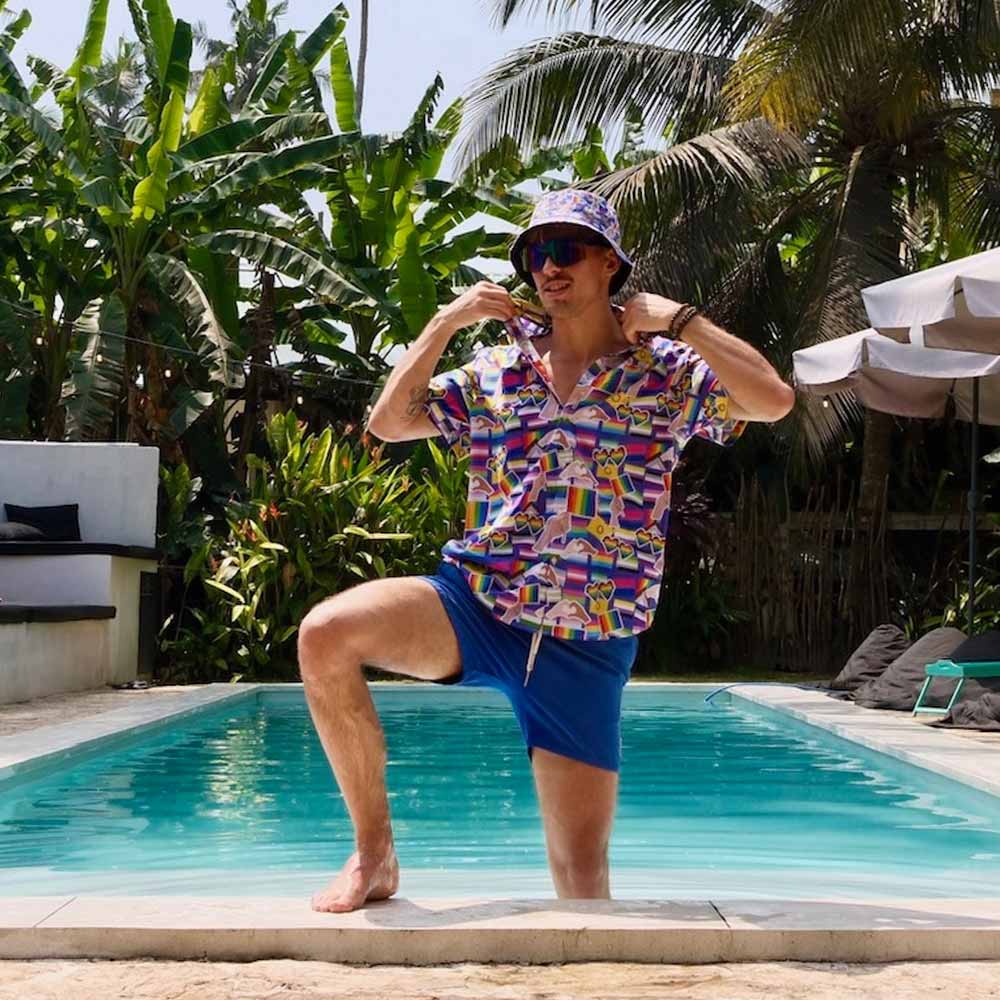 A man wearing an LGBTQ Pride Shirt, blue shorts, and a matching bucket hat poses with one knee up by a swimming pool, surrounded by lush tropical plants and palm trees.