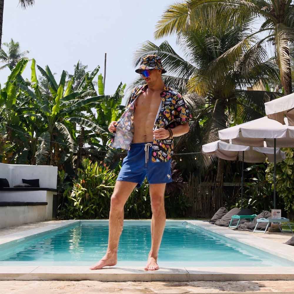 A man in the Wanna Pizza This Shirt from Bad Shirt Club, blue shorts, and a matching bucket hat stands confidently by a pool lined with palm trees, holding his shirt open to reveal his bare chest.