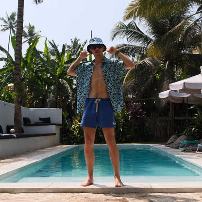 Wearing a Turquoise Tempest Leopard Print Bucket Hat, sunglasses, a patterned shirt, and blue swim trunks, a man stands confidently by the pool amid tropical plants and palm trees under the sunny sky.