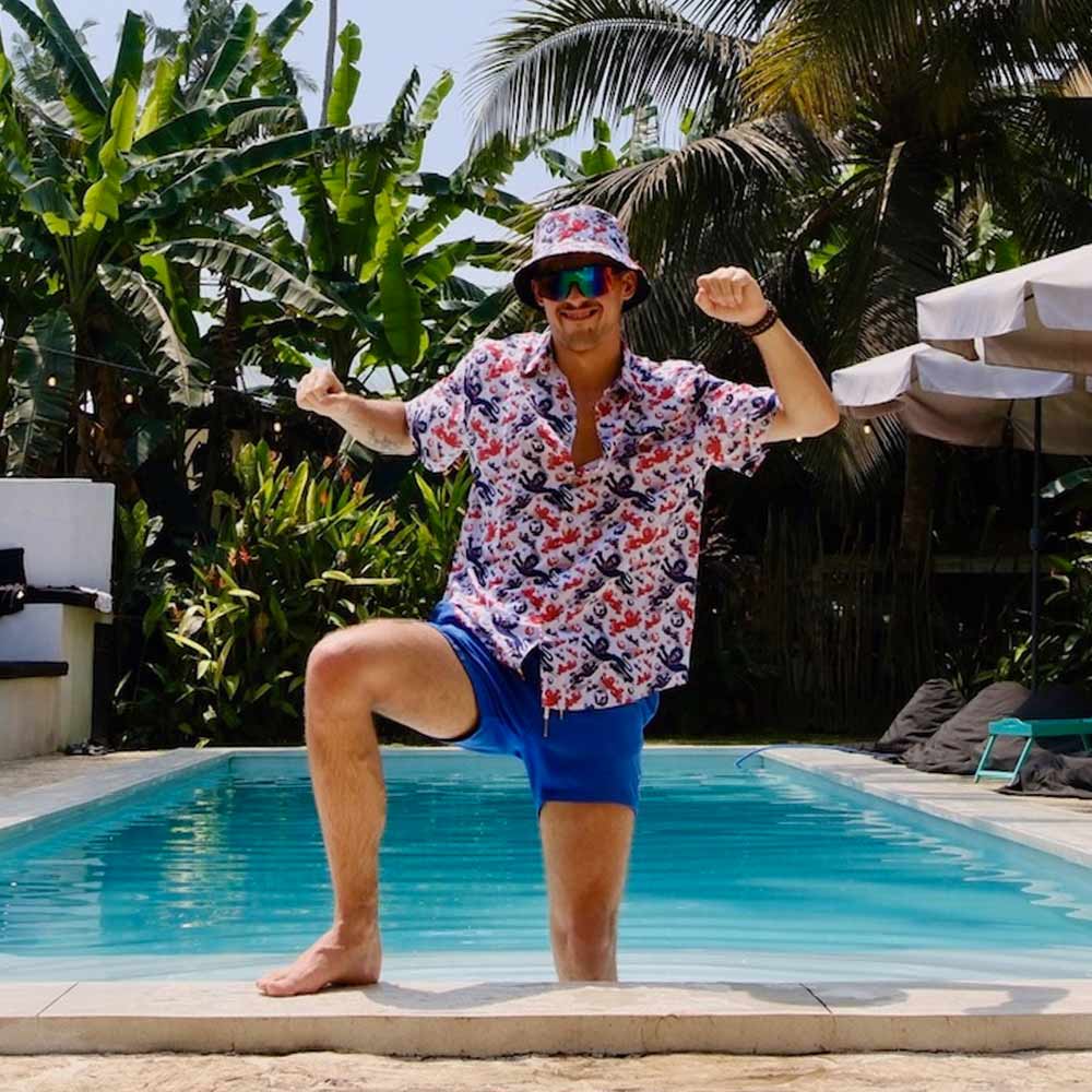 A man wearing the England Football Hawaiian Shirt, blue shorts, a bucket hat, and sunglasses poses by a pool amid lush tropical plants and palm trees.