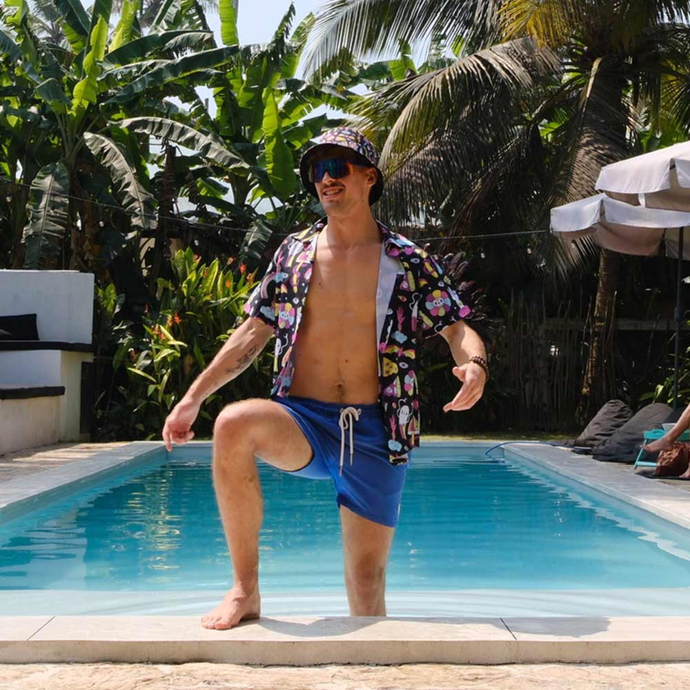 Wearing the Shroom Shirt, blue shorts, and sunglasses, a man stands with one foot on a pool’s edge. Tropical plants and palm trees frame the sunny scene as he enjoys the day.