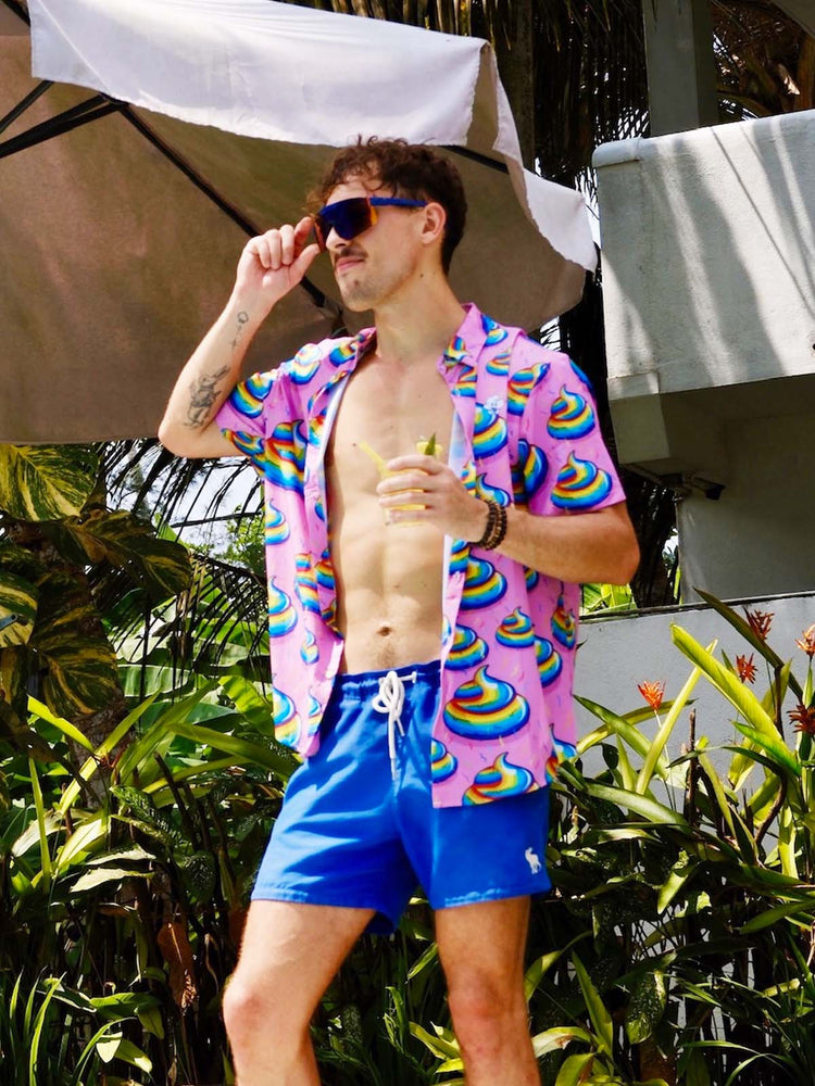A man in sunglasses and a bright pink shit shirt with colourful swirl unicorn poo pattern stands outside by tropical plants, holding a drink and wearing blue swim trunks, with a large umbrella and building in the background.