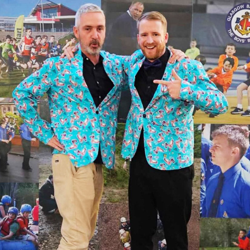 Two men pose together, smiling and wearing matching bright blue jackets with a colorful pattern. The background features a collage of photos showing people, outdoor activities, and a round Glasgow Boys’ Brigade logo.