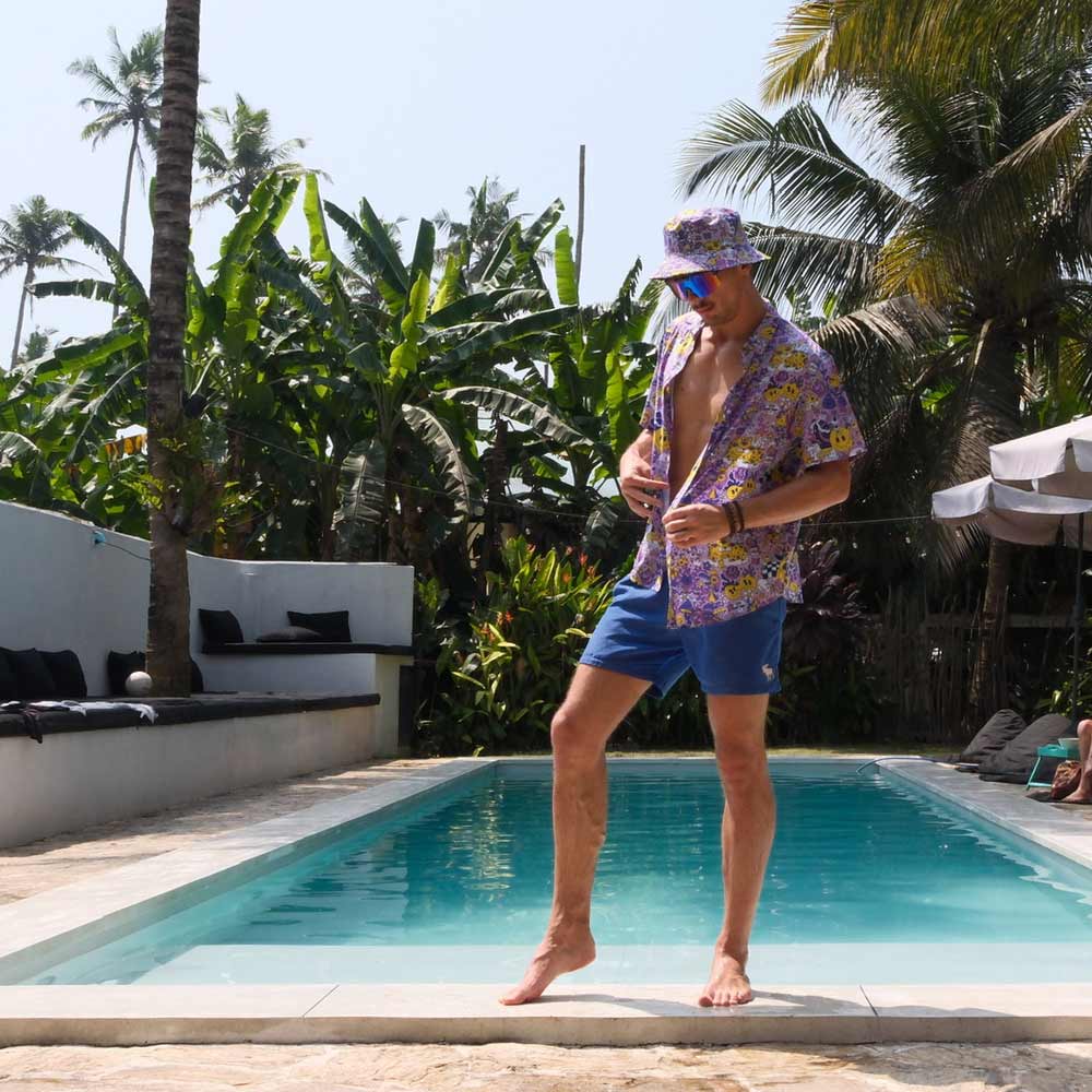 Wearing a Magical Psychedelic Shirt, blue shorts, and a bucket hat, a man stands at the edge of a swimming pool, one foot raised as if stepping onto the water, surrounded by lush tropical plants and palm trees.