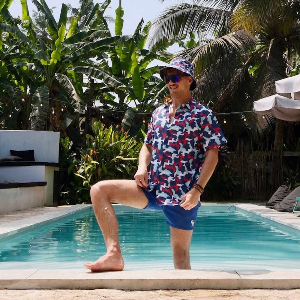 A man poses by a pool in a Combat2Coffee Army Camo Shirt and blue swim shorts, raising mental health awareness for veterans amid tropical plants and palm trees under a sunny sky.