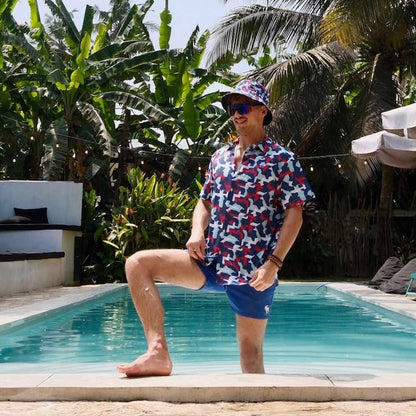 A man poses by a pool in a Combat2Coffee Army Camo Shirt and blue swim shorts, raising mental health awareness for veterans amid tropical plants and palm trees under a sunny sky.