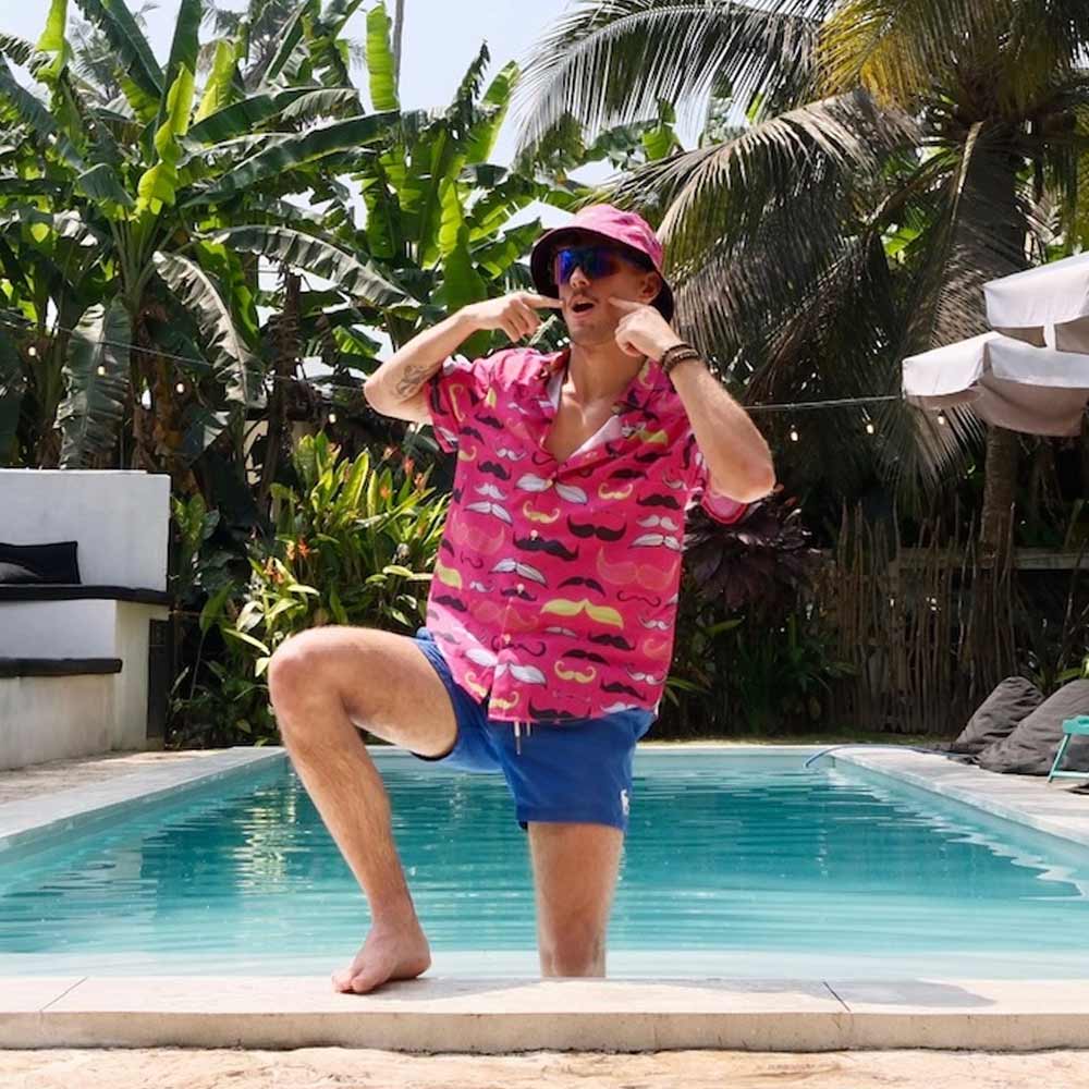 A man wearing sunglasses, a colorful hat, blue shorts, and the Movember Shirt with pink mustaches poses playfully by a pool amid tropical plants.