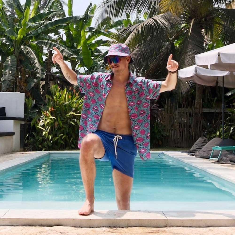 A man in the Leopard Kiss Bad Shirt with bold feline prints, blue shorts, and a bucket hat poses poolside with one knee up, pointing skyward. Palm trees and lounge chairs complete the ultimate summer vibe.