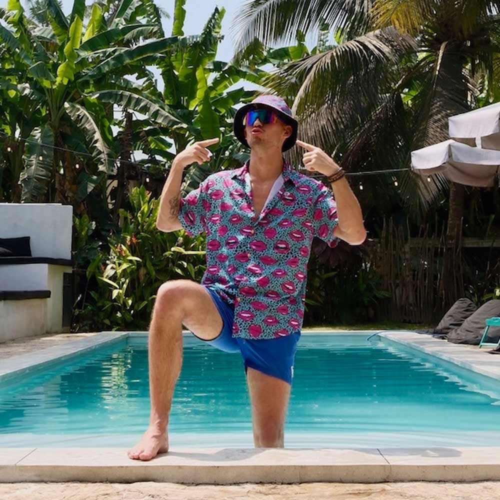 Wearing the Leopard Kiss Bad Shirt, blue shorts, and a sunhat, a man kneels confidently by the pool and points at himself—a bold look in tropical surroundings with palm trees and lush plants.