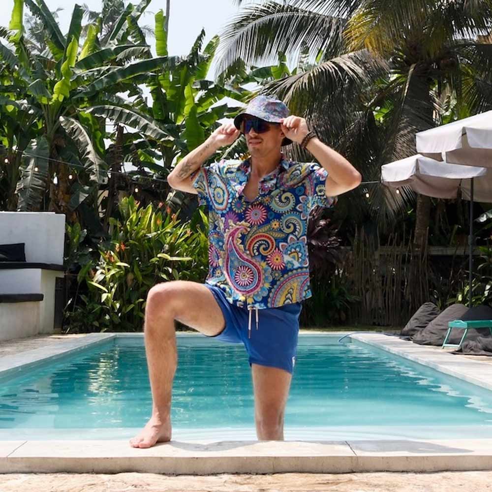 A man wearing the Psychedelic Explorer Bucket Hat, a vibrant shirt, and blue shorts poses poolside with one knee raised, smiling as he holds his hat. Tropical plants and palm trees fill the background.
