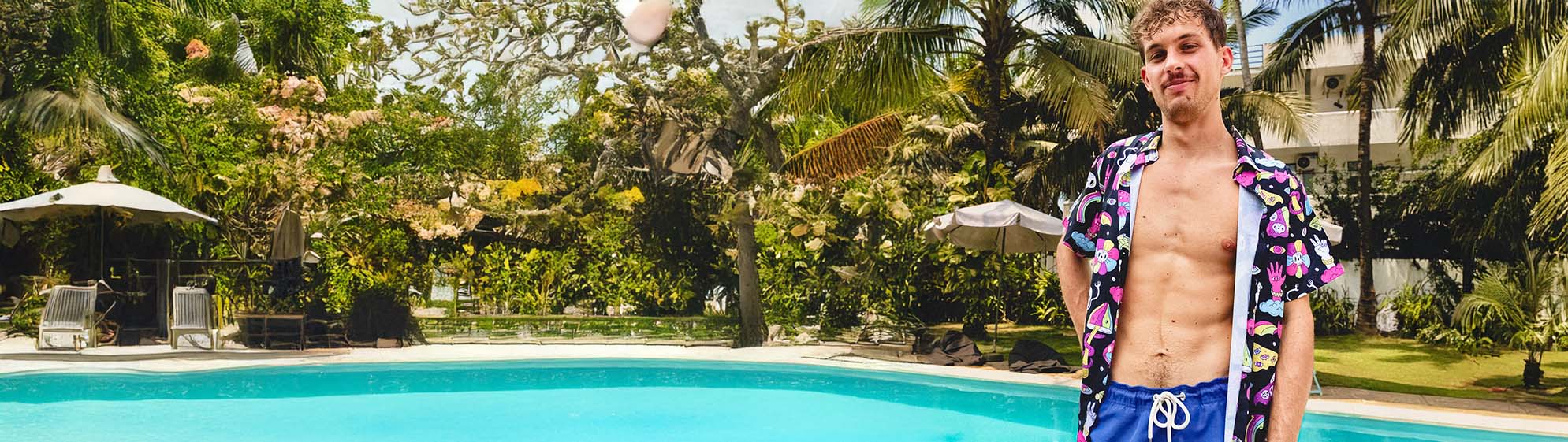 A young man wearing a patterned open shit shirt and blue swim trunks stands by a bright blue outdoor pool, surrounded by palm trees and greenery on a sunny day.
