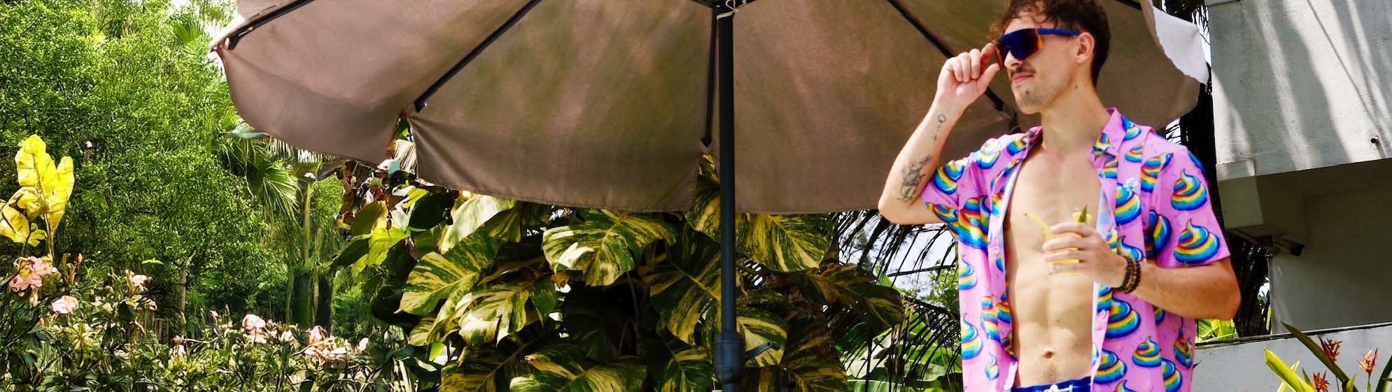 A man wearing a colourful shit shirt, swim trunks, and sunglasses stands under a large outdoor umbrella, holding a drink. He is outside near lush tropical plants and a building, enjoying a sunny day.
