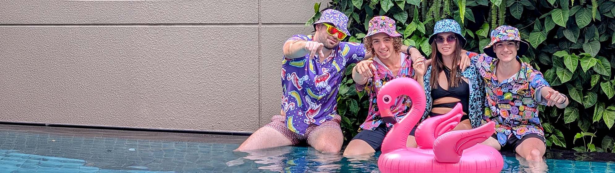 Four people in colourful matching shit shirt outfits and bucket hats pose by a pool with a large pink inflatable flamingo. Green foliage and a beige wall are in the background.