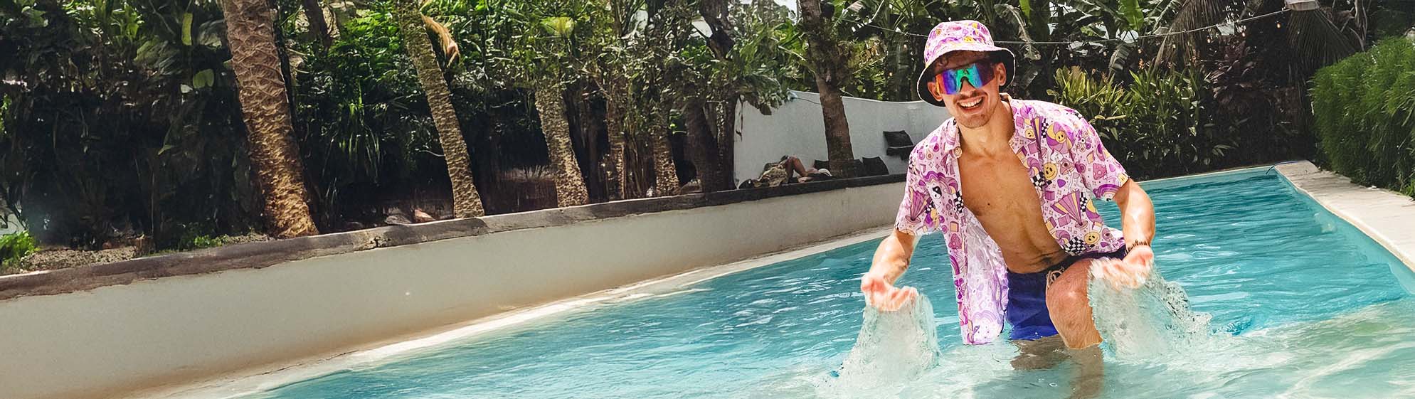 A man wearing a colorful bucket hat, sunglasses, and a pink patterned shirt splashes in a narrow outdoor pool surrounded by lush tropical plants and palm trees.