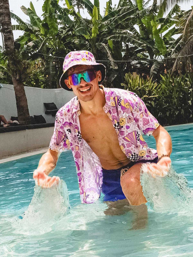 A smiling man in a colorful shirt, matching bucket hat, and sunglasses stands in a pool splashing water, surrounded by palm trees and tropical plants.